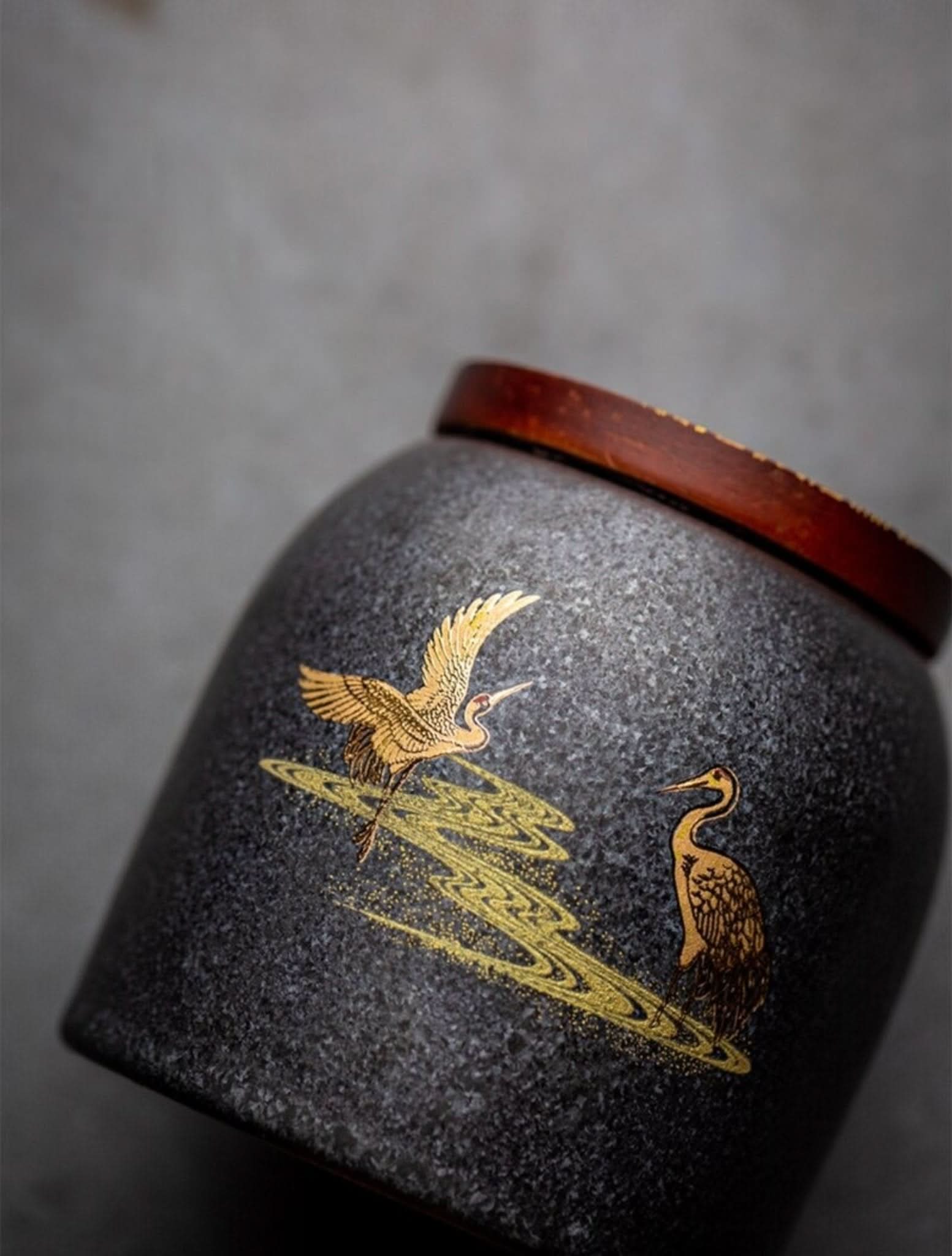 Retro black ceramic tea canister with gold cranes and a wooden lid on a grey surface.